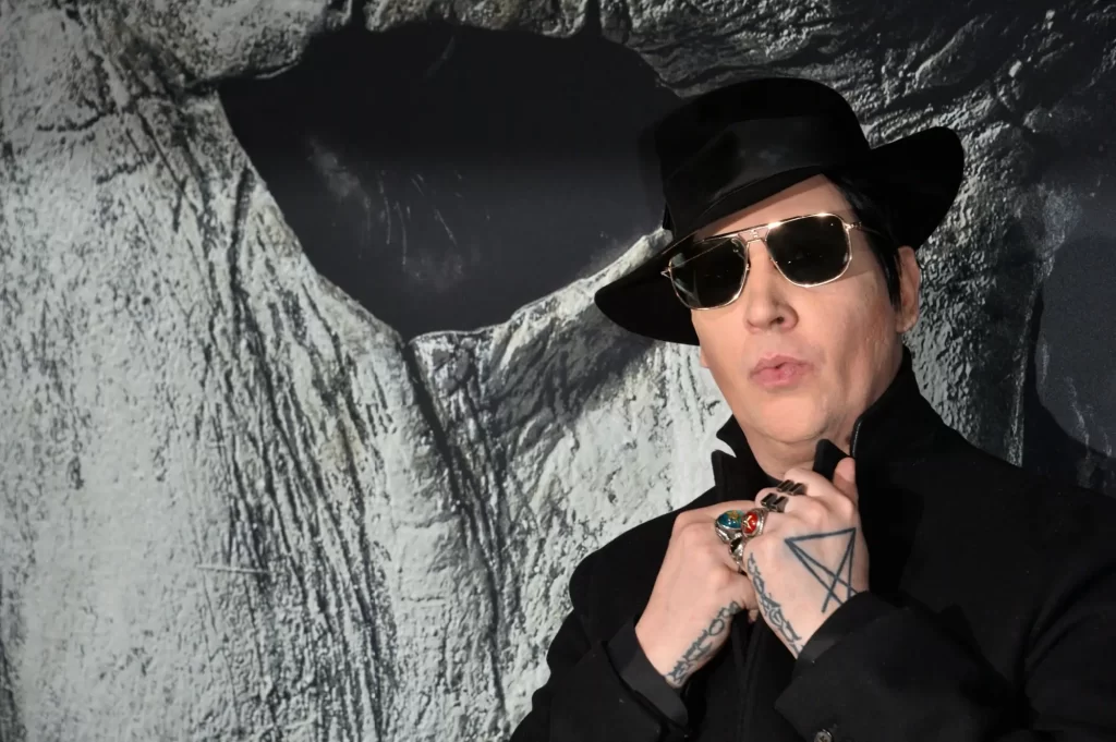 Marilyn Manson v černém outfitu, s výraznými tetováními na rukou, tmavými brýlemi a kloboukem. 