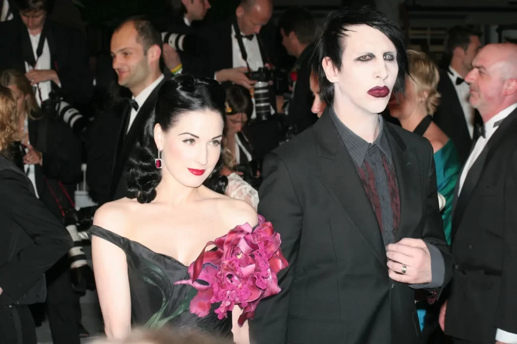 Marilyn Manson v tmavém obleku s typickým bledým make-upem, černými vlasy a rudou rtěnkou. Po jeho boku kráčí burleskní hvězda Dita Von Teese v elegantních černých šatech s výraznými šperky a květinou v ruce.