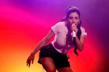 MAria na koncertu s mikrofonem v ruce, bílé tričko a černé kraťasy.