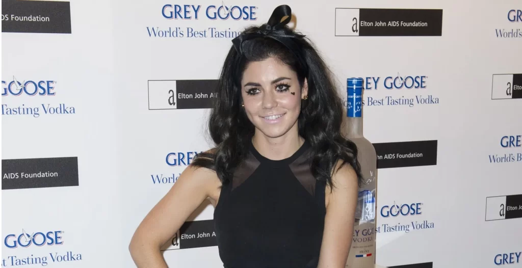 - VIPshow Marina Diamandis s tmavými, objemně zvlněnými vlasy, ozdobenými černou mašlí. Má na sobě černé šaty bez rukávů s průhledným detailem v oblasti dekoltu.