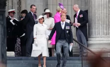 Meghan v bílých šatech a Harry ve smokingu sestupují po schodech. Za nimi královská rodina