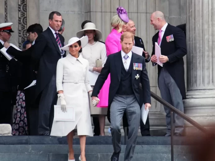 Meghan v bílých šatech a Harry ve smokingu sestupují po schodech. Za nimi královská rodina