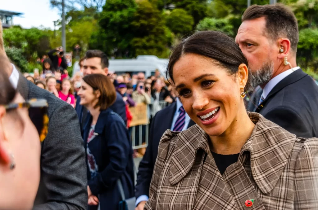 Meghan Markle při veřejném setkání s občany. Má na sobě elegantní kostkovaný kabát s červeným květem na klopě a působí uvolněně a přátelsky.
