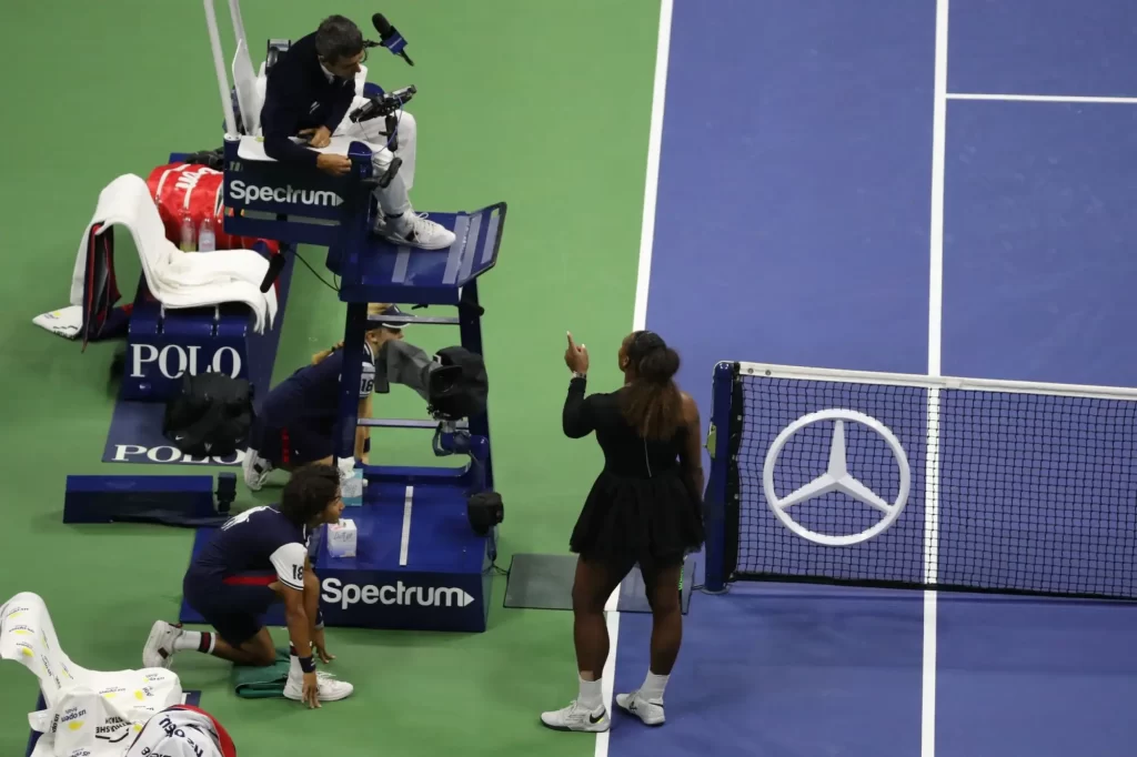 Serena, oblečená v černém sportovním outfitu, stojí u rozhodčího na umpiru a energicky gestikuluje.