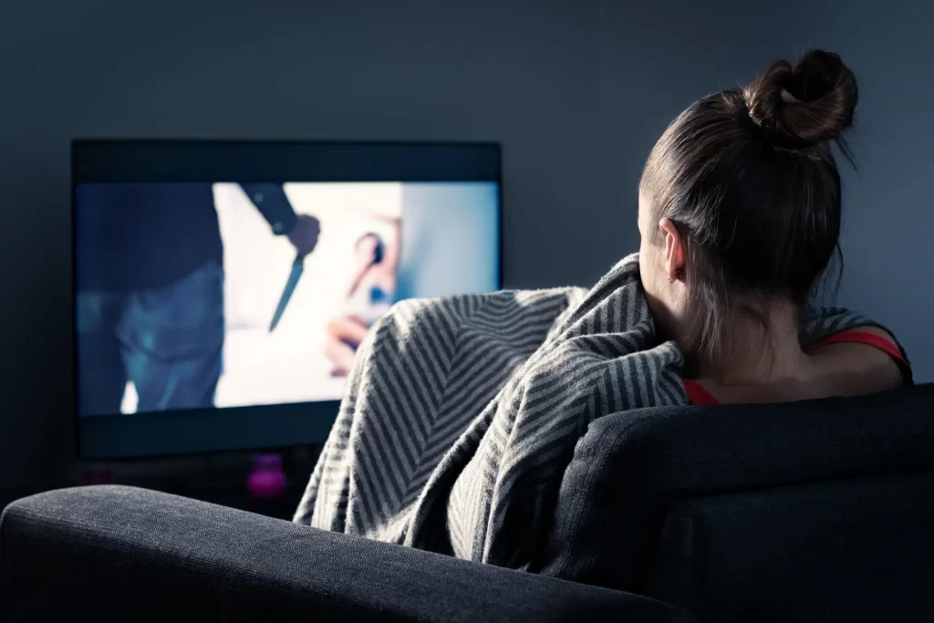Žena sedící na pohovce zabalená v šedé pruhované dece sleduje napínavou true crime scénu.