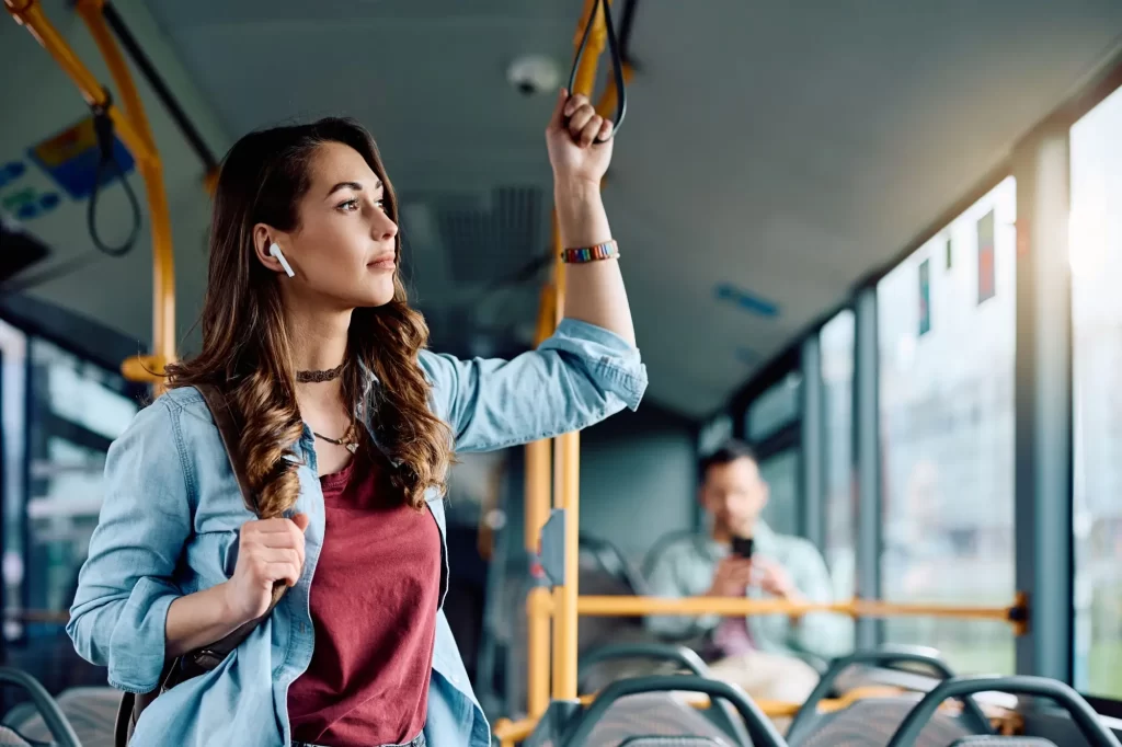 Mladá žena cestuje městským autobusem, drží se madla a poslouchá něco ve sluchátkách.