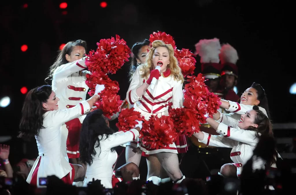 Madonna během živého vystoupení na stadionu, obklopena tanečnicemi v bílých uniformách s červenými detaily. 