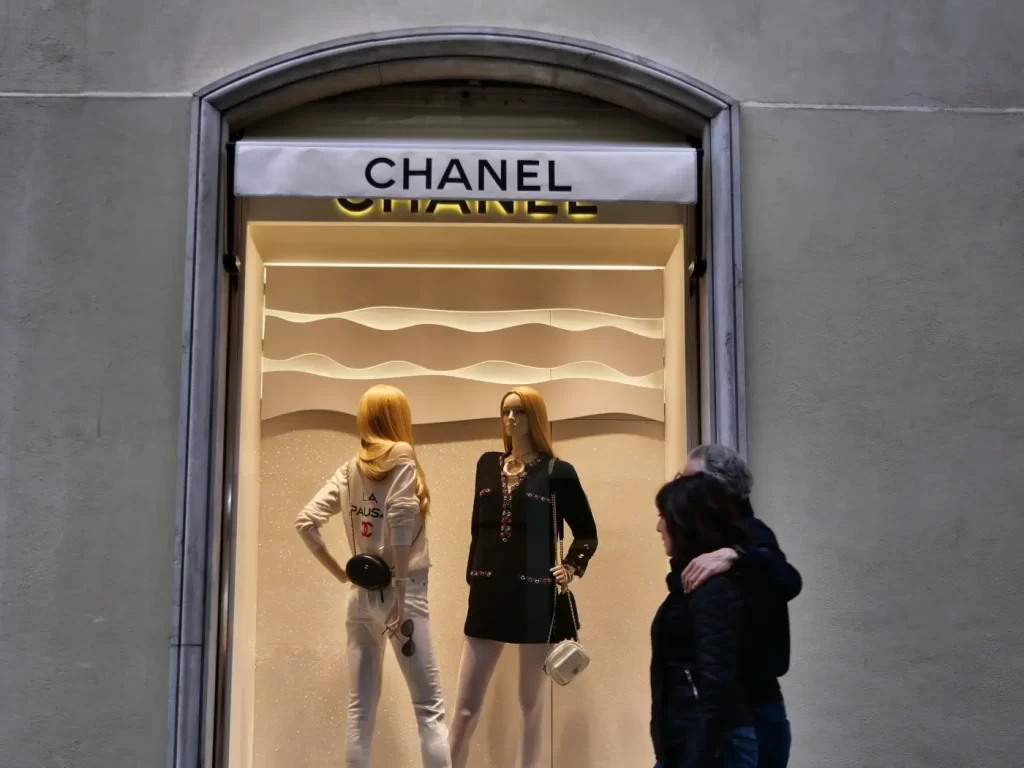 Prosklená výloha butiku Chanel s figurínami v moderních outfitech, u které prochází dvojice lidí