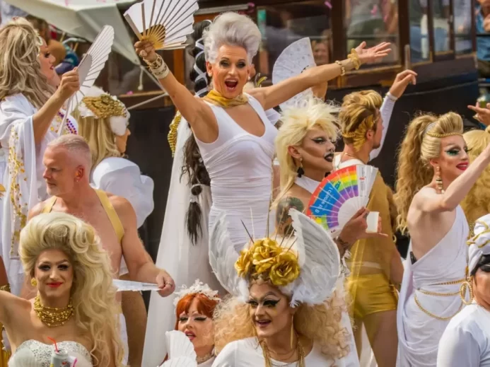 Skupina drag performerek v bílých a zlatých kostýmech slaví během průvodu. Výrazné make-upy, vějíře a výrazy nadšení