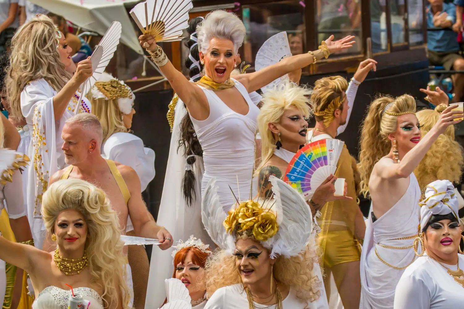 Skupina drag performerek v bílých a zlatých kostýmech slaví během průvodu. Výrazné make-upy, vějíře a výrazy nadšení