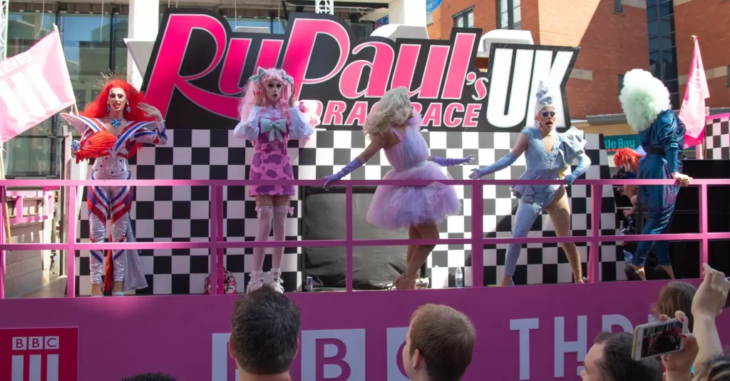 Pódium s účastnicemi RuPaul’s Drag Race UK během veřejné akce. Barevné kostýmy, extravagantní paruky a pózy před nadšeným publikem ukazují, jak silně pronikl drag do popkultury.