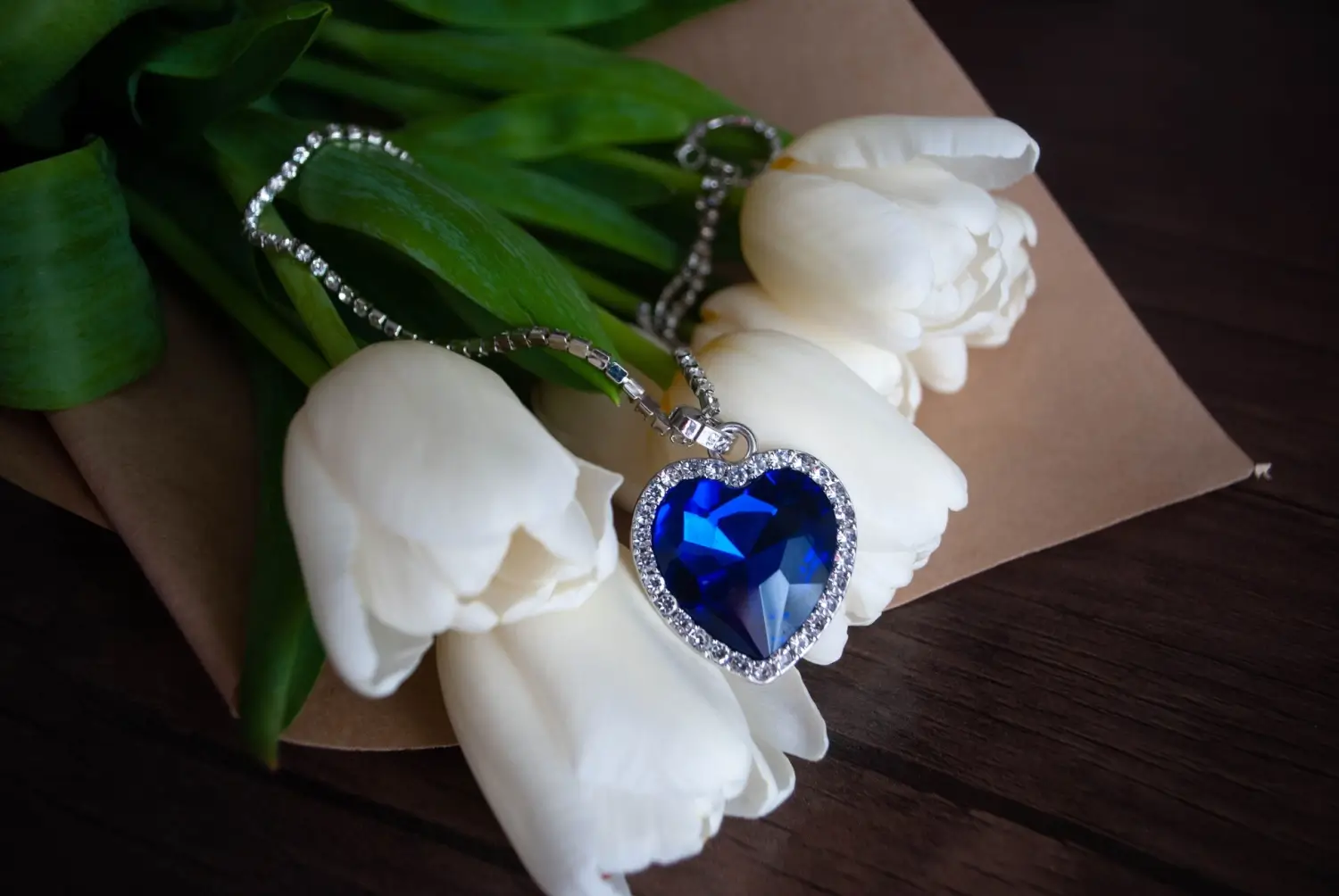Modrý srdcový náhrdelník posázený diamanty položený na bílých tulipánech