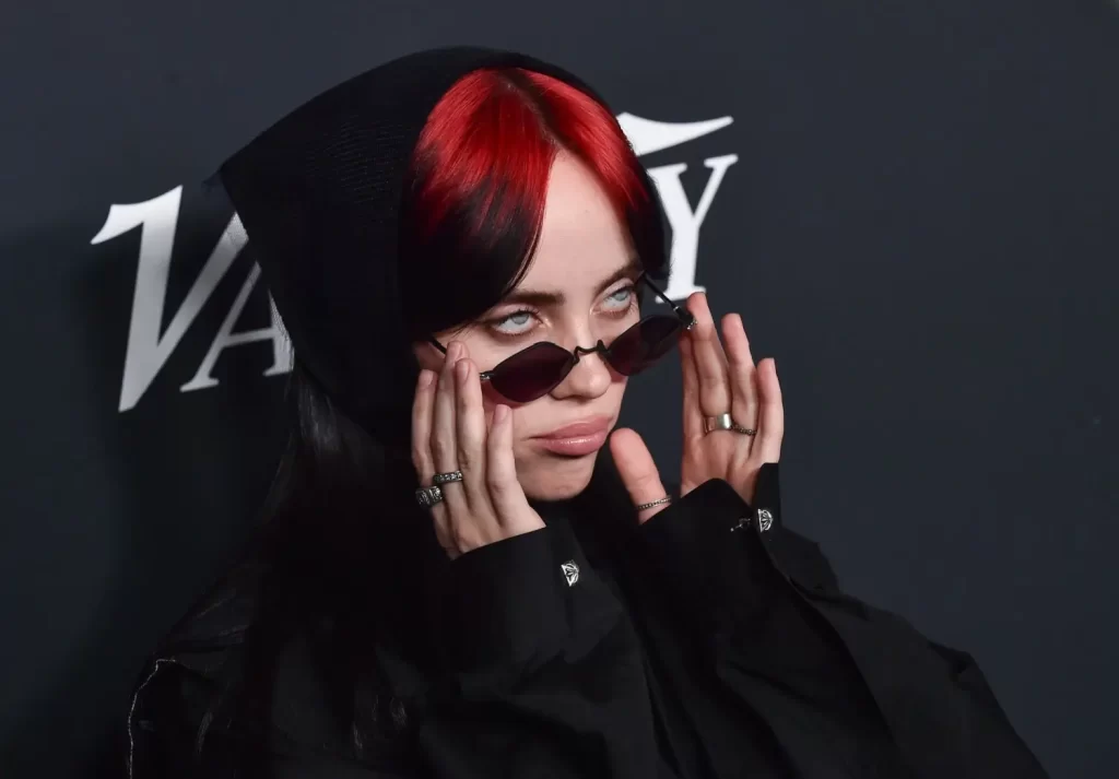 Billie Eilish v černém oblečení a s červeno-černými vlasy si nasazuje sluneční brýle