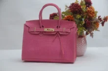 Růžová krokodýlí kabelka Birkin s výrazným zlatým kováním, postavená vedle vázy s květinami