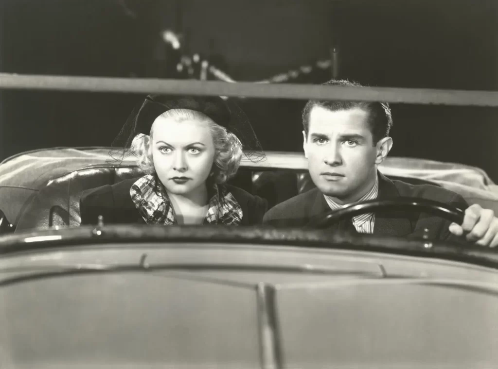 Černobílá filmová scéna zachycuje ženu a muže v klasickém automobilu, oba s vážným výrazem a pohledem upřeným vpřed. 