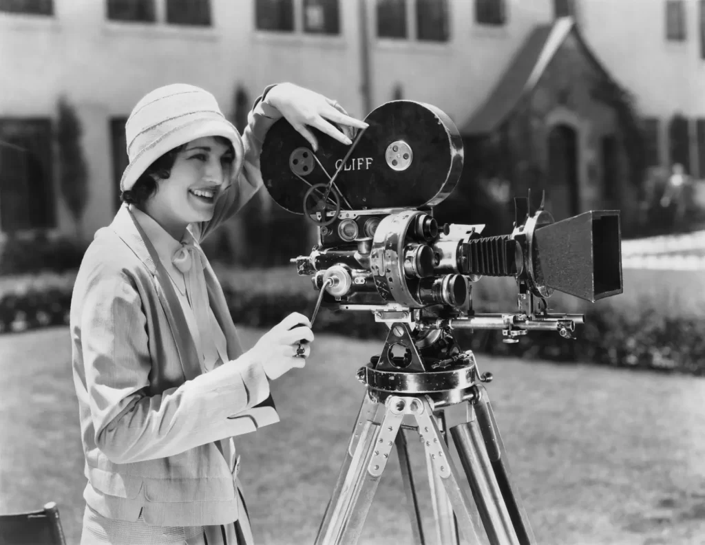 Usměvavá žena ve stylovém klobouku a dobovém oblečení stojí venku u klasické filmové kamery na stativu.