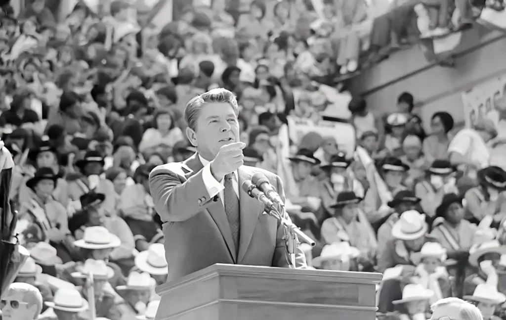 Ronald Reagan v obleku za řečnickým pultem, hovoří do mikrofonů a gestikuluje sevřenou pěstí.