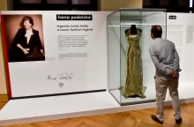 Muž si prohlíží zlaté večerní šaty ve vitríně na výstavě věnované Haně Podolské, ikonické české módní návrhářce