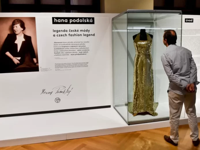 Muž si prohlíží zlaté večerní šaty ve vitríně na výstavě věnované Haně Podolské, ikonické české módní návrhářce