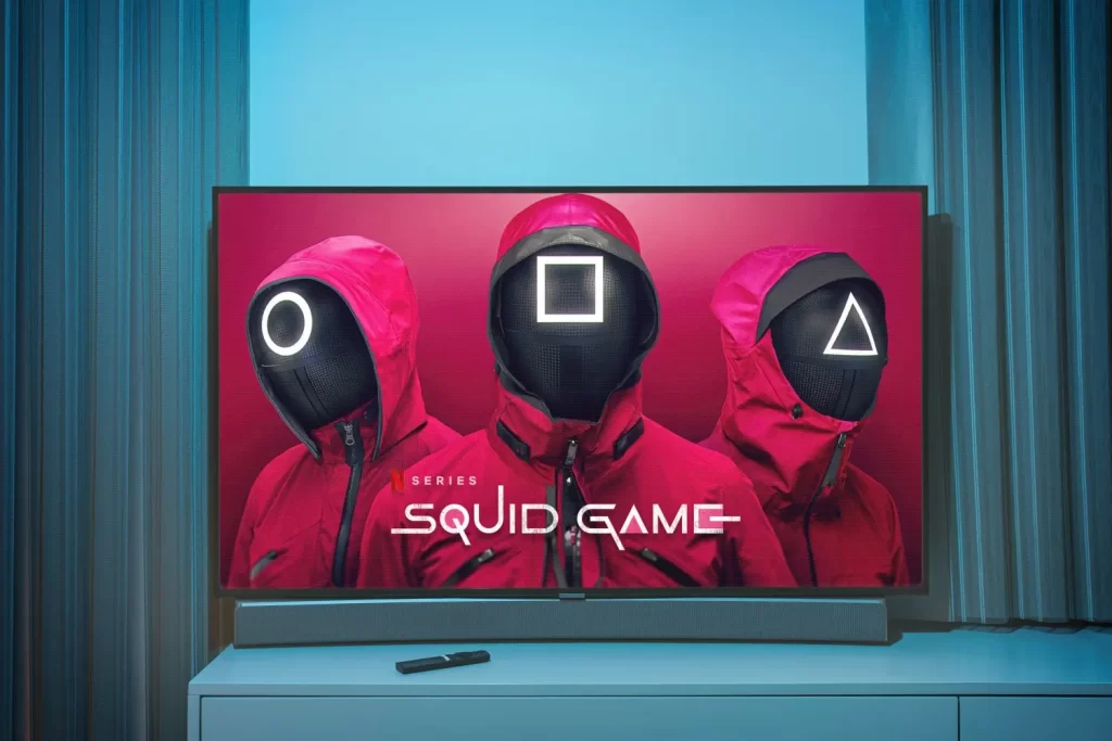 Televizní obrazovka se záběrem ze seriálu Squid Game, ikonické postavy ve červených kostýmech s geometrickými tvary na maskách