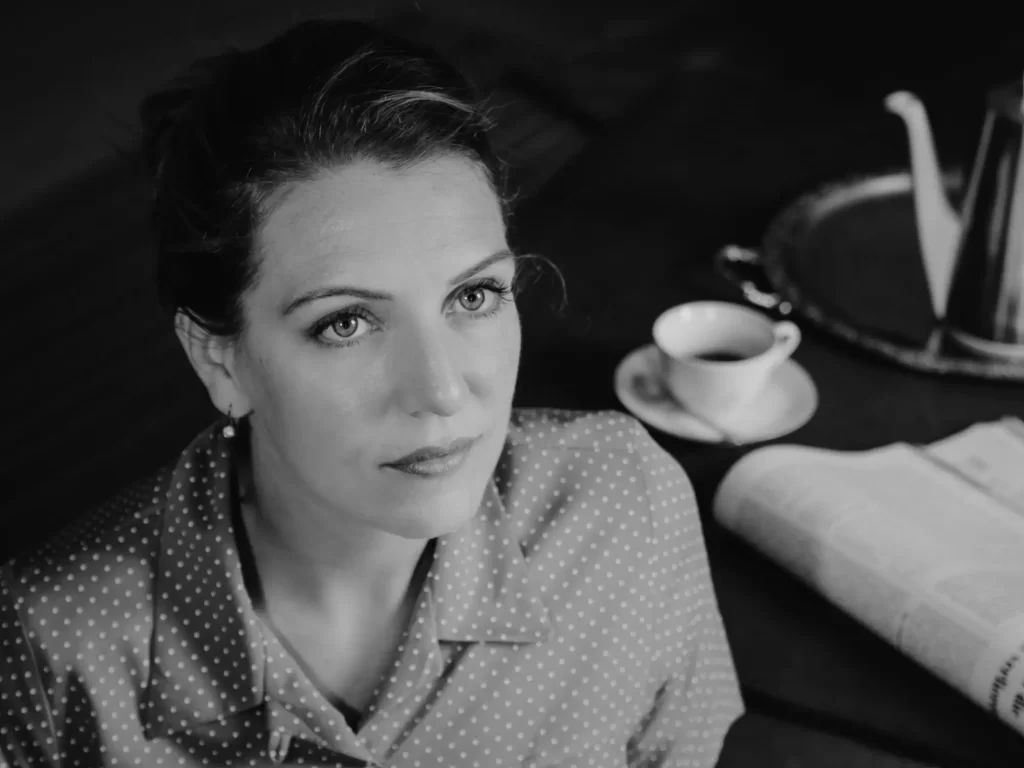 Žena v retro stylu, sedící u stolu s šálkem kávy, konvičkou a novinami.