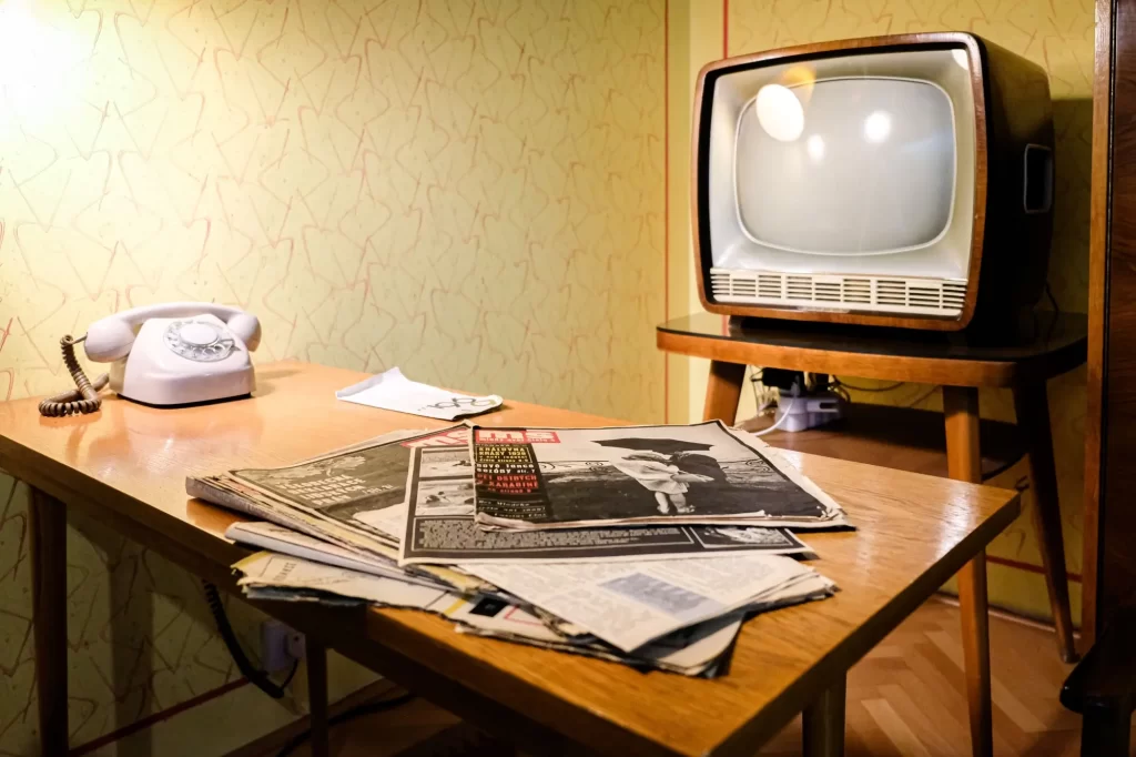 Na dřevěném stole leží staré noviny nebo filmové magazíny, vedle nich klasický otočný telefon.