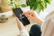 ohled na ruce člověka, který spouští aplikaci TikTok na chytrém telefonu, v pozadí je hrnek kávy a zelené pokojové rostliny