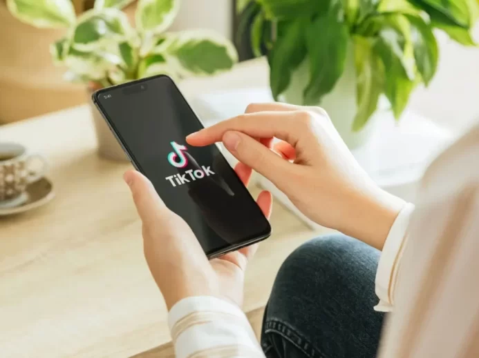 ohled na ruce člověka, který spouští aplikaci TikTok na chytrém telefonu, v pozadí je hrnek kávy a zelené pokojové rostliny