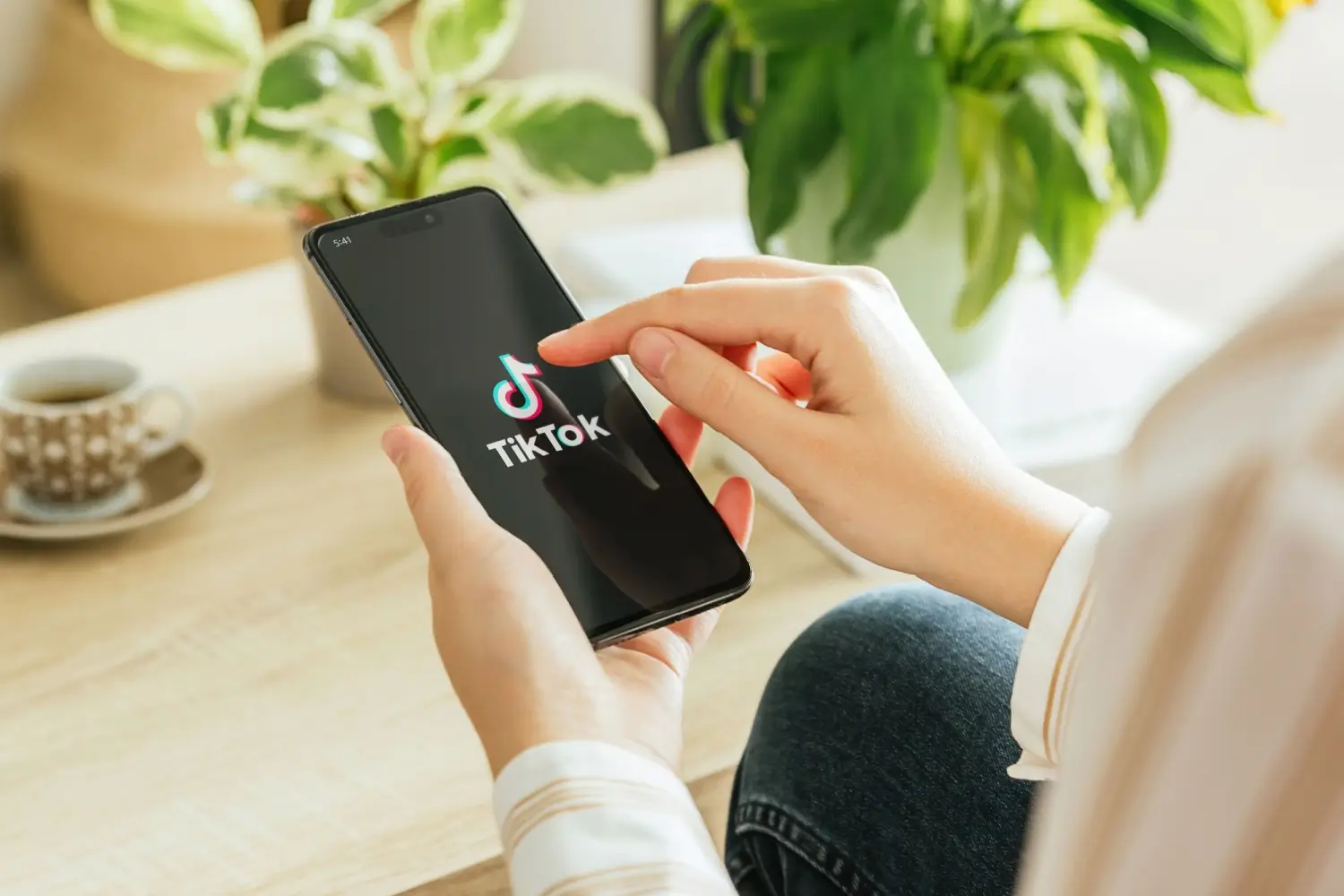 ohled na ruce člověka, který spouští aplikaci TikTok na chytrém telefonu, v pozadí je hrnek kávy a zelené pokojové rostliny