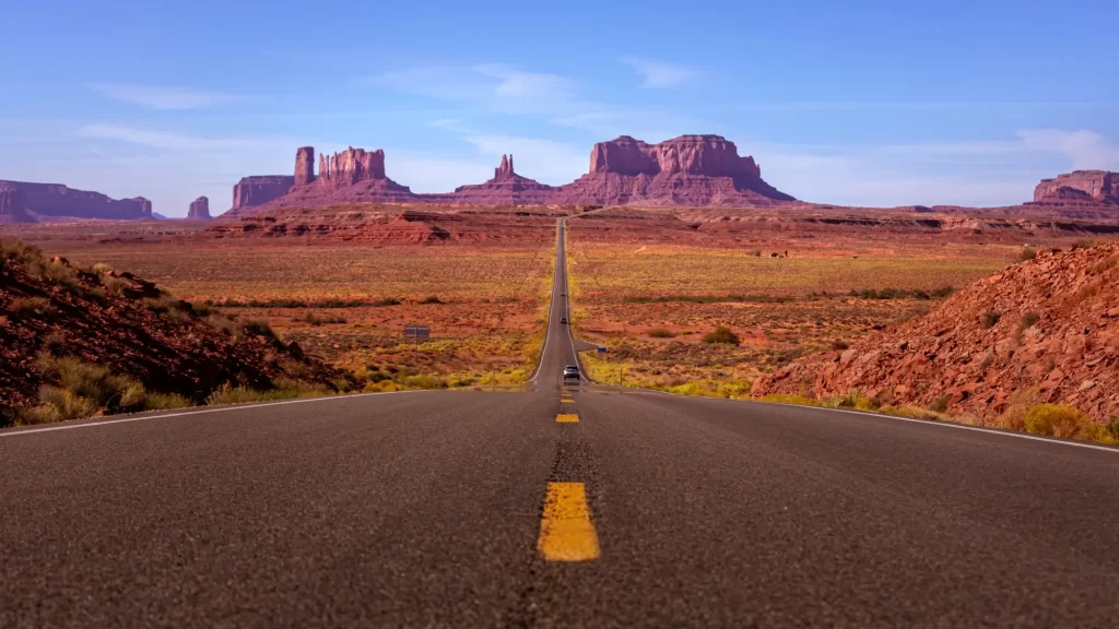 Na obrázku je dlouhá rovná silnice směřující k monumentálním skalním útvarům v oblasti Monument Valley v USA.