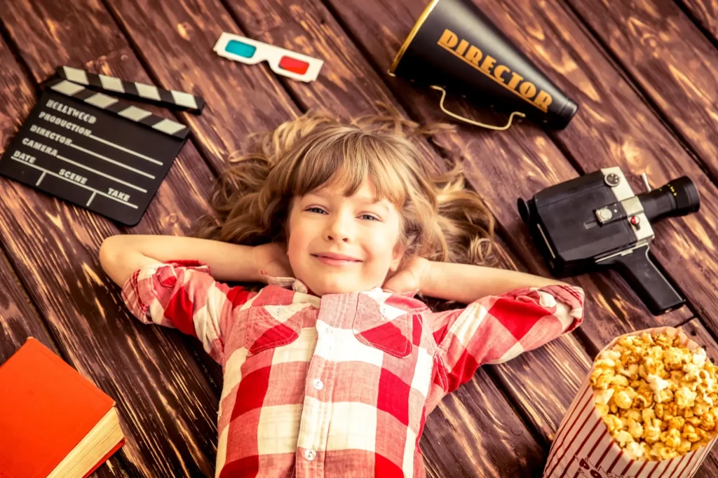 Chlapec v kostkované košili na dřevěné podlaze, kolem něj jsou filmové rekvizity jako klapka, kamera, megafon, 3D brýle a popcorn.
