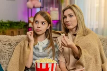 Matka s dcerou společně sledují televizi s popcornem a přikryté dekou