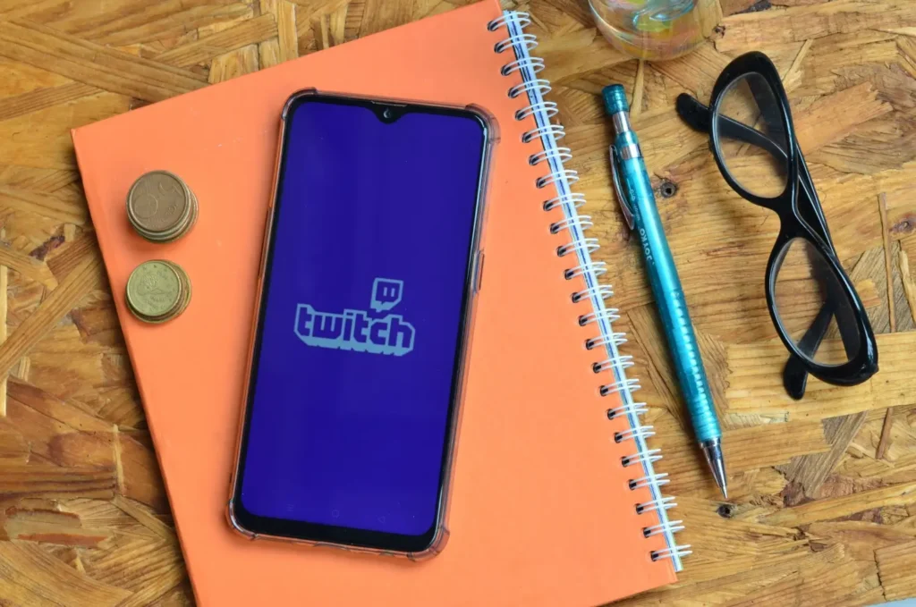 Smartphone s otevřenou aplikací Twitch ležící na sešitě, vedle brýlí, tužky a mincí