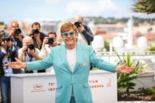 Elton John během fotografování na Filmovém festivalu v Cannes v roce 2019. Má na sobě tyrkysově modrý oblek s broží na klopě a bílé tričko. Jeho ikonické modré brýle s třpytivými obroučkami.