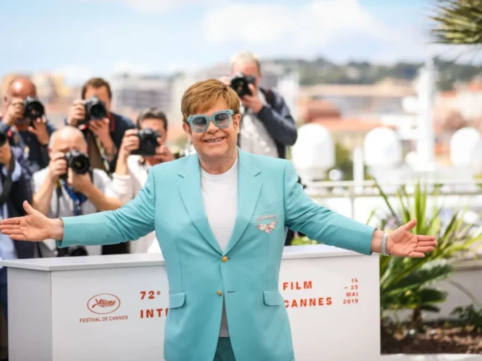 Elton John během fotografování na Filmovém festivalu v Cannes v roce 2019. Má na sobě tyrkysově modrý oblek s broží na klopě a bílé tričko. Jeho ikonické modré brýle s třpytivými obroučkami.