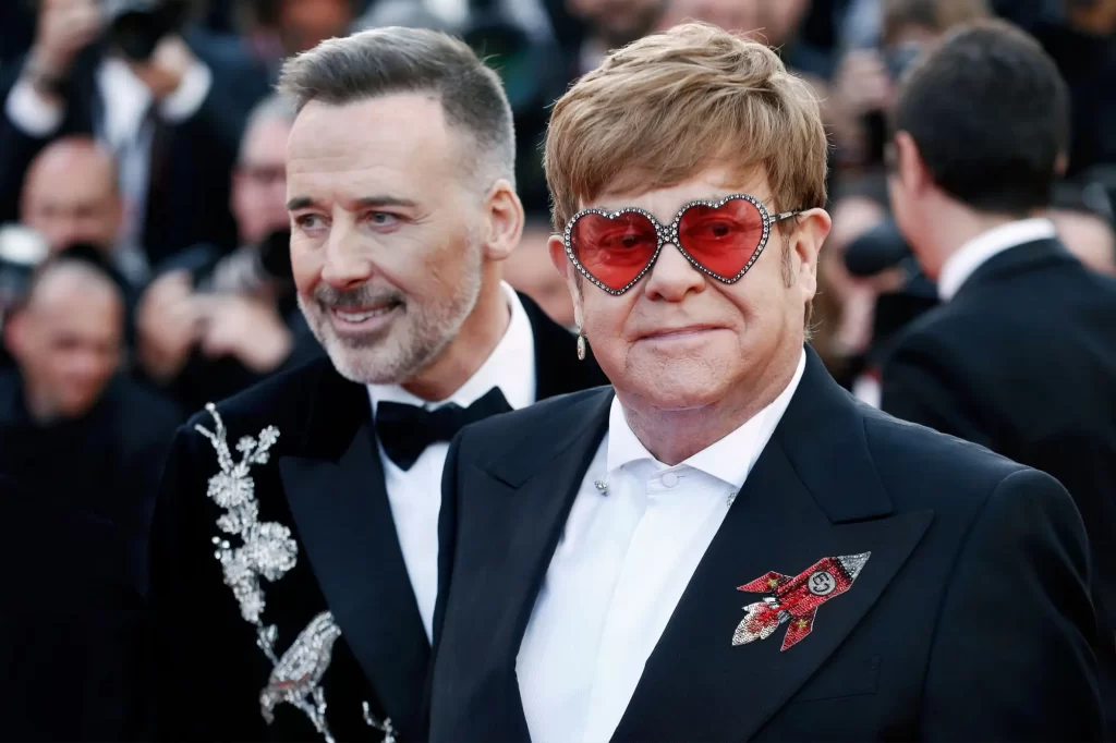 - VIPshow Elton John na červeném koberci v elegantním černém saku s broží ve tvaru rakety a výraznými brýlemi ve tvaru červených srdcí zdobenými kamínky.