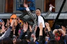 Jihokorejský zpěvák PSY, známý díky megahitu Gangnam Style, který stojí nad davem fanoušků s rukama vztyčenýma do vzduchu.