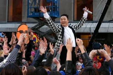 Jihokorejský zpěvák PSY, známý díky megahitu Gangnam Style, který stojí nad davem fanoušků s rukama vztyčenýma do vzduchu.