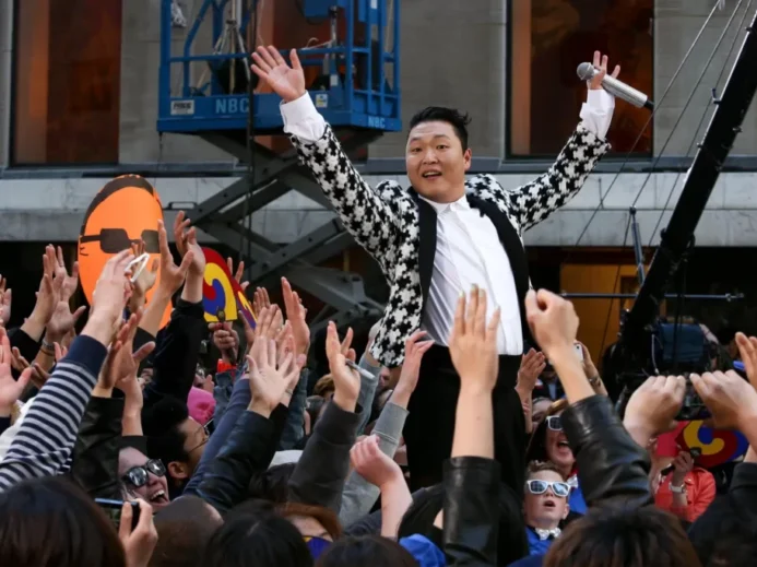 Jihokorejský zpěvák PSY, známý díky megahitu Gangnam Style, který stojí nad davem fanoušků s rukama vztyčenýma do vzduchu.