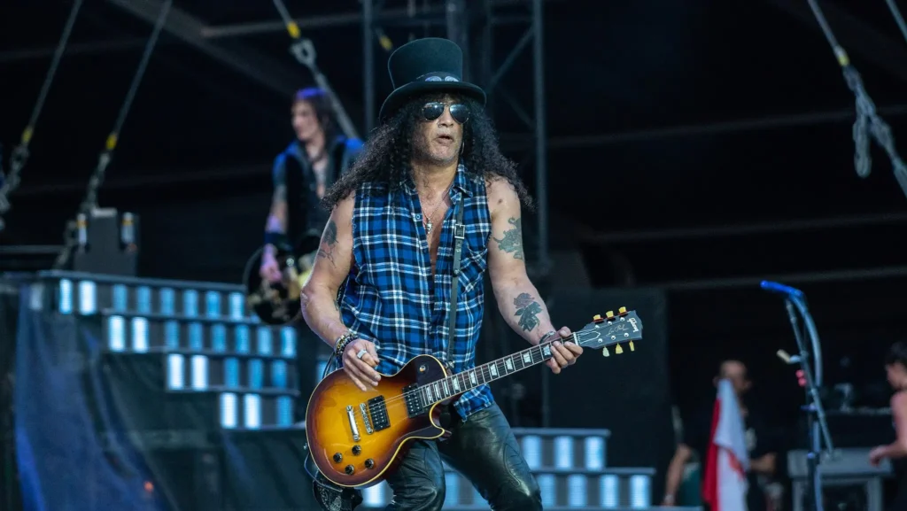 - VIPshow Kytarista Slash z Guns N’ Roses hraje na pódiu v klobouku a kostkované košili