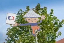 Ilustrace postavičky Večerníčka s papírovou čepicí a logem České televize před zelenými stromy