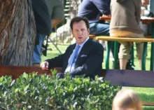 Americký herec James Woods sedí na lavičce v parku v tmavém obleku