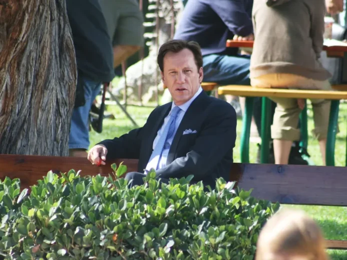 Americký herec James Woods sedí na lavičce v parku v tmavém obleku