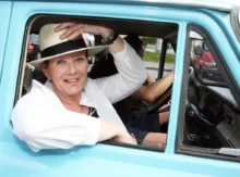 Žena si drží klobouk, sedí v automobilu