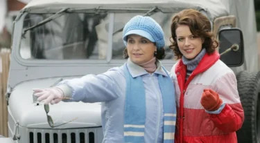 Two women in winter clothing stand next to a vintage jeep; one points ahead while holding sunglasses, the other smiles beside her.