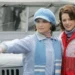 Two women in winter clothing stand next to a vintage jeep; one points ahead while holding sunglasses, the other smiles beside her.
