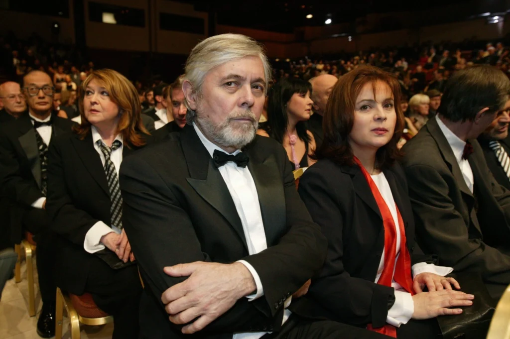 Dva lidé sedí v publiku, Muž se mračí a má založené ruce, žena se mírně usmívá.