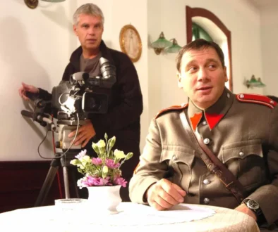 Muž v četnické uniformě sedí u stolu a za ním stojí další muž