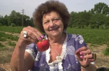 Starší žena oblečená v šatech stojí venku a v ruce drží velkou jahodu