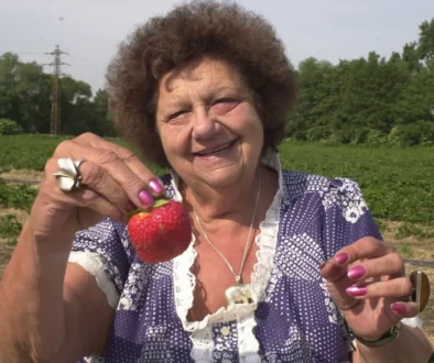 Starší žena oblečená v šatech stojí venku a v ruce drží velkou jahodu